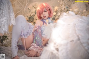 A woman with pink hair sitting on a bed holding a mirror.