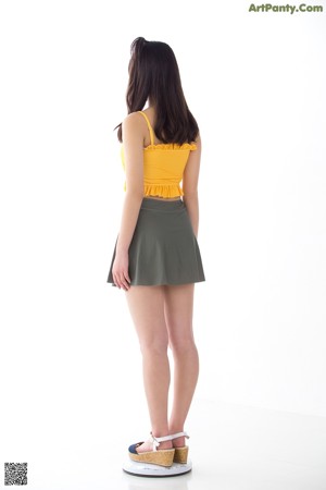 A woman in a yellow top and green skirt leaning against a wall.